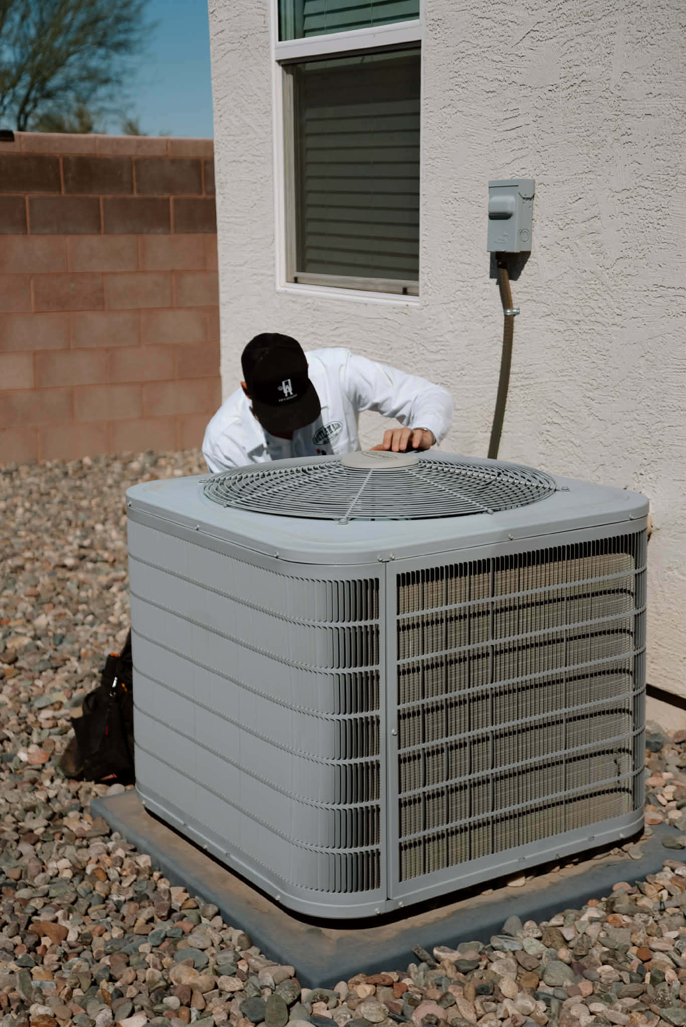 Outlaw Air technician checking the top of an outdoor AC condenser beside a stucco home
