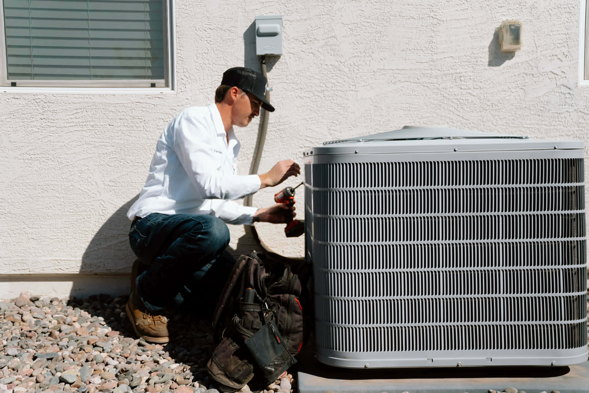 Outlaw Air technician kneeling beside an outdoor AC condenser next to a stucco home