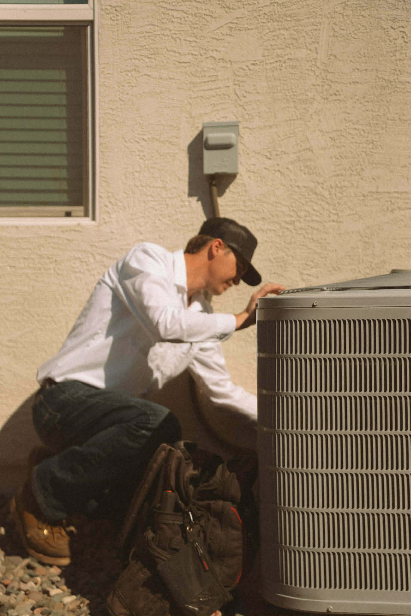 Outlaw Air technician crouched beside an outdoor AC condenser against a stucco wall