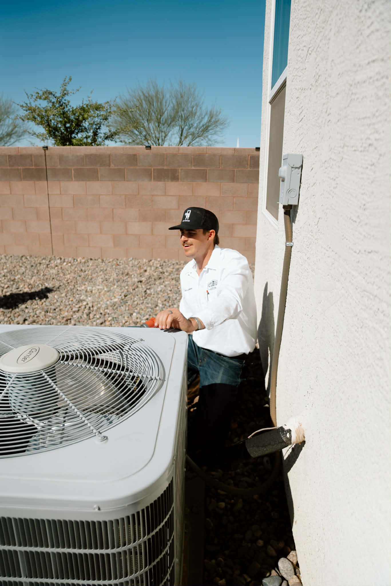 Outlaw Air technician kneeling behind an outdoor AC condenser in a gravel side yard